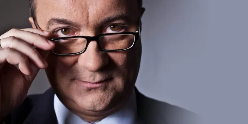 Zbliżenie mężczyzny w okularach, który jedną ręką poprawia szkła i patrzy w obiektyw na jasnoszarym tle.