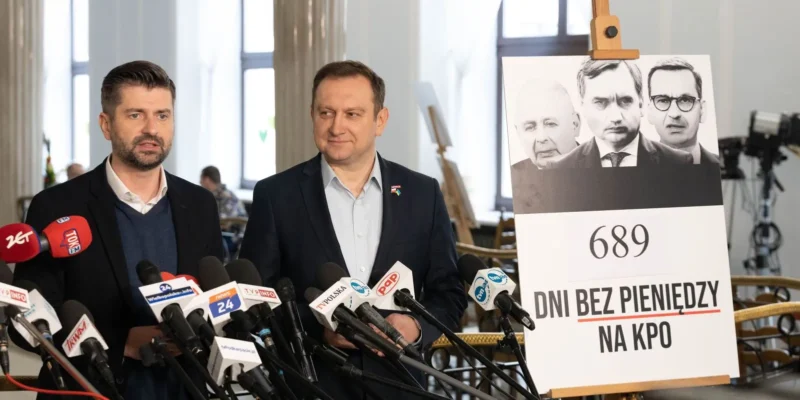 Dwóch mężczyzn w garniturach przemawia podczas konferencji prasowej, otoczonych mikrofonami, obok planszy z napisem „689 dni bez pieniędzy na KPO” i czarno-białymi portretami.