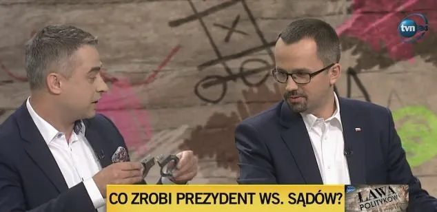 Dwóch mężczyzn w marynarkach rozmawia w studiu telewizyjnym podczas programu publicystycznego, jeden trzyma kajdanki, a u dołu ekranu widoczny jest napis "CO ZROBI PREZYDENT WS. SĄDÓW?".