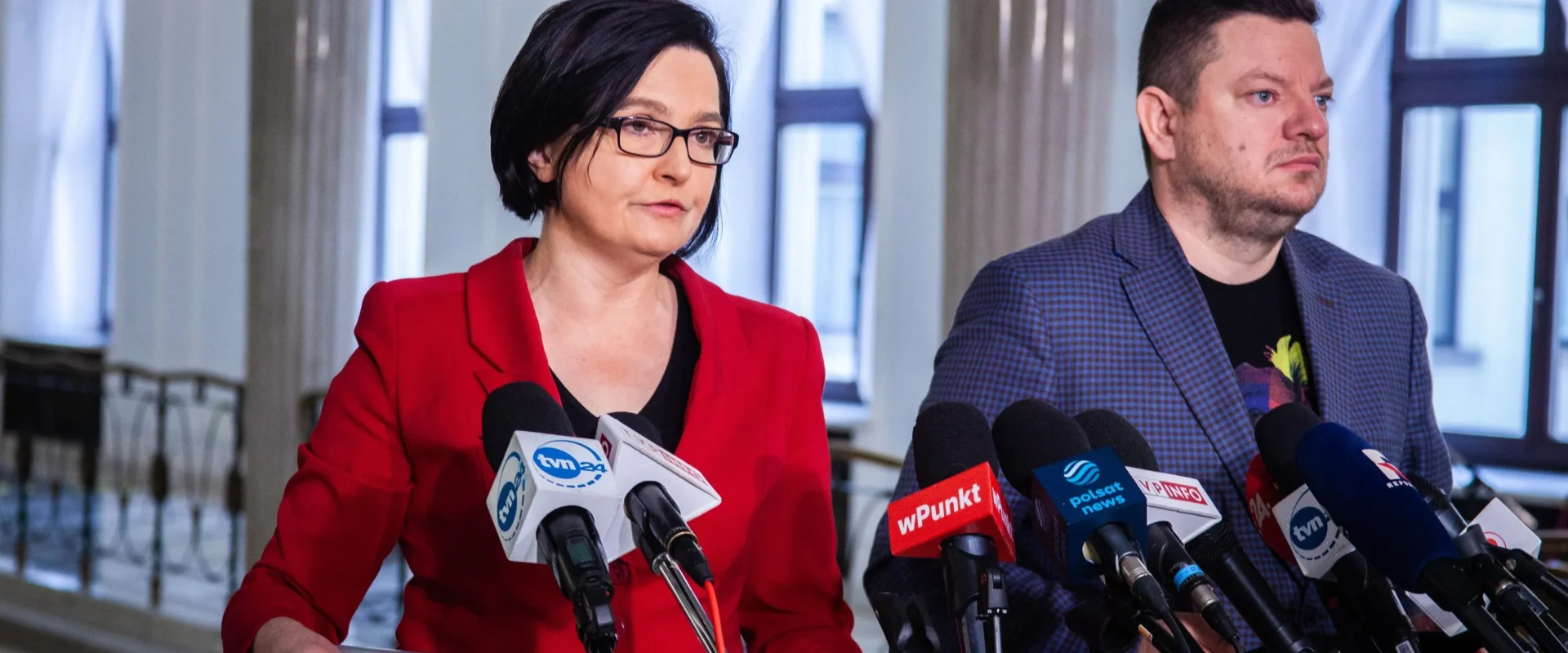 Na zdjęciu widać dwóch mówców stojących przy dźwigni z mikrofonami, w otoczeniu jasnego wnętrza.