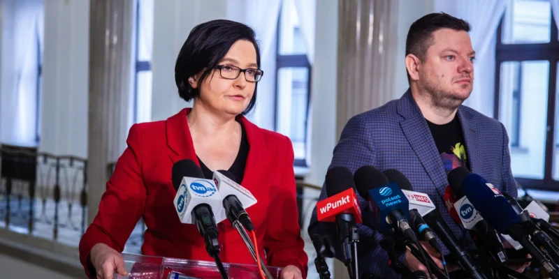 Na zdjęciu widać dwóch mówców stojących przy dźwigni z mikrofonami, w otoczeniu jasnego wnętrza.