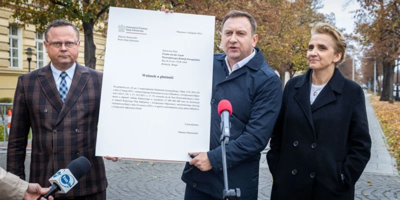 Na zdjęciu trzej urzędnicy, w tym jeden trzymający dokument, prezentują wniosek o płatność związany z funduszami europejskimi.