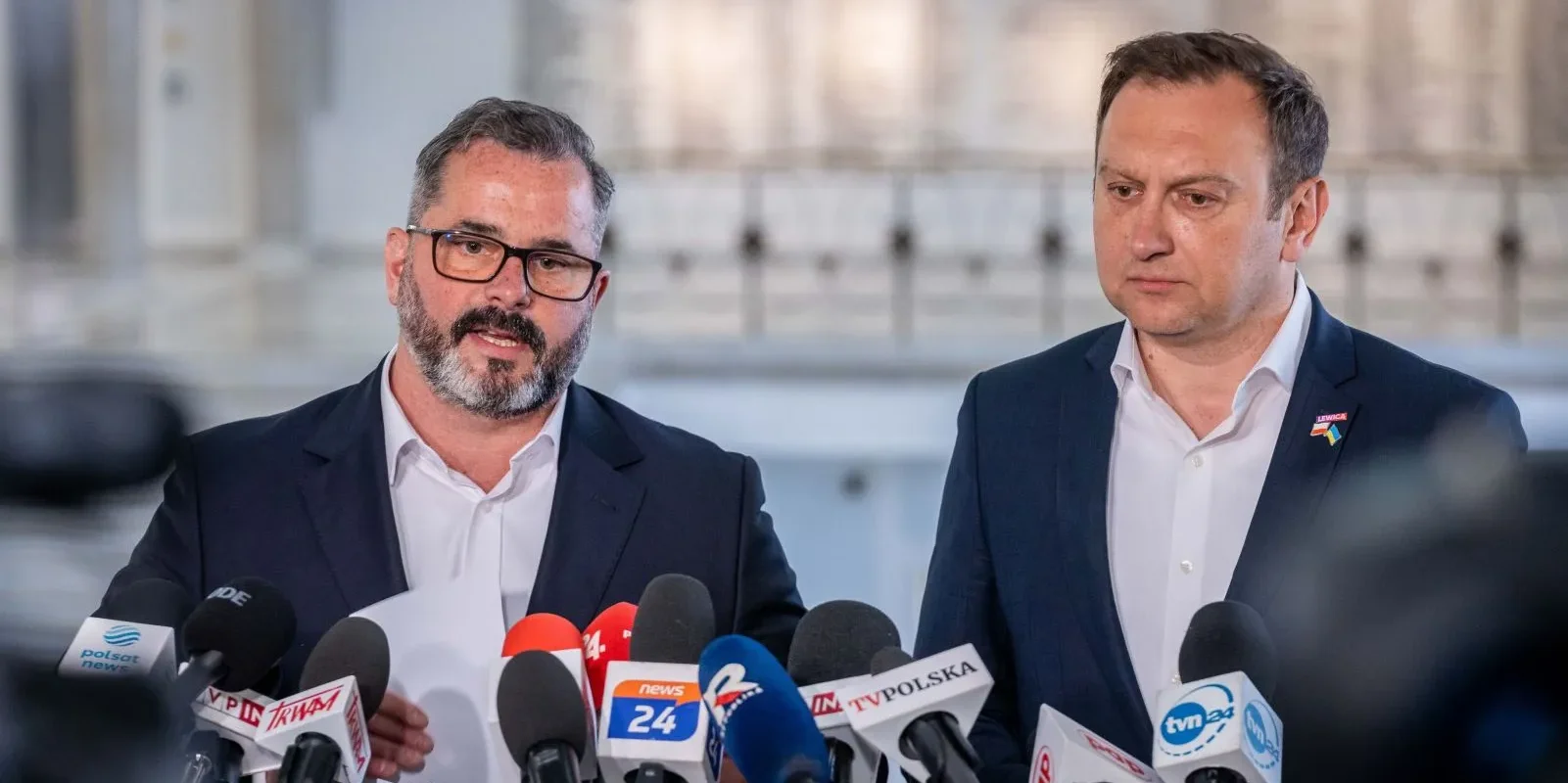 Na zdjęciu widać dwóch mężczyzn podczas konferencji prasowej, stojących przed mikrofonami z różnych mediów w jasnym, przestronnym otoczeniu.