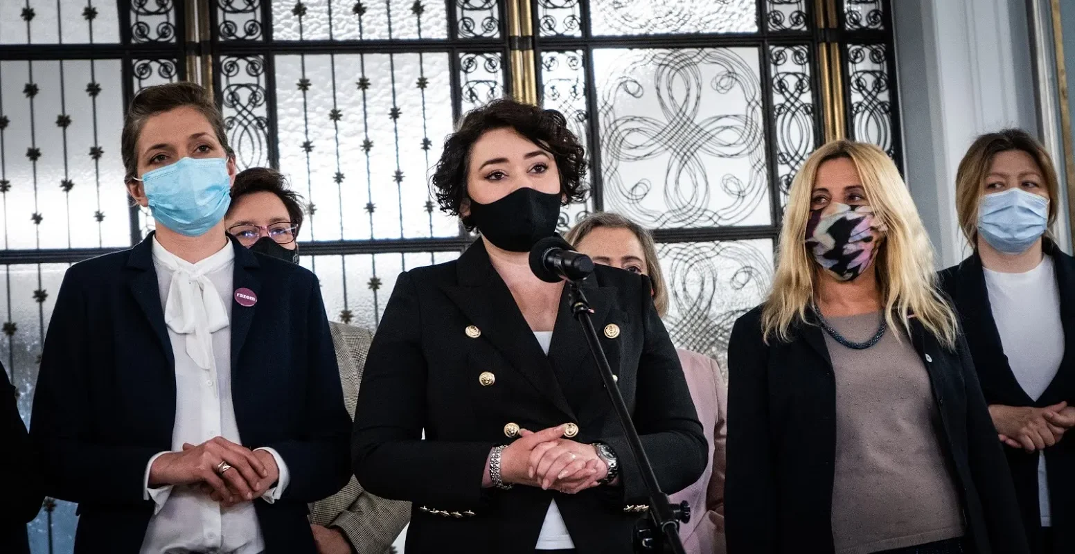 Kobieta w czarnym garniturze przemawia do mikrofonu podczas konferencji prasowej, otoczona innymi kobietami w maseczkach, a w tle widać ozdobne metalowe okno.