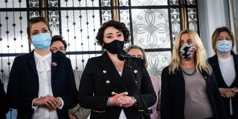 Kobieta w czarnym garniturze przemawia do mikrofonu podczas konferencji prasowej, otoczona innymi kobietami w maseczkach, a w tle widać ozdobne metalowe okno.