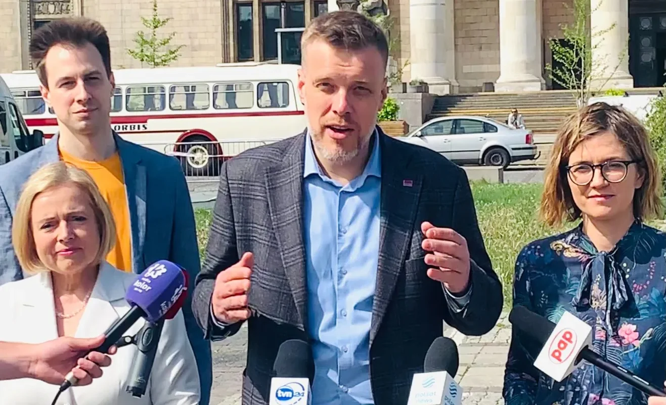 Grupa osób prowadzi konferencję prasową na zewnątrz budynku z kolumnami; mężczyzna w kratkowanej marynarce gestykuluje do mediów, otoczony dziennikarzami z mikrofonami.