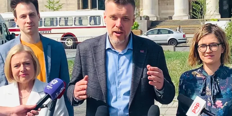 Grupa osób prowadzi konferencję prasową na zewnątrz budynku z kolumnami; mężczyzna w kratkowanej marynarce gestykuluje do mediów, otoczony dziennikarzami z mikrofonami.