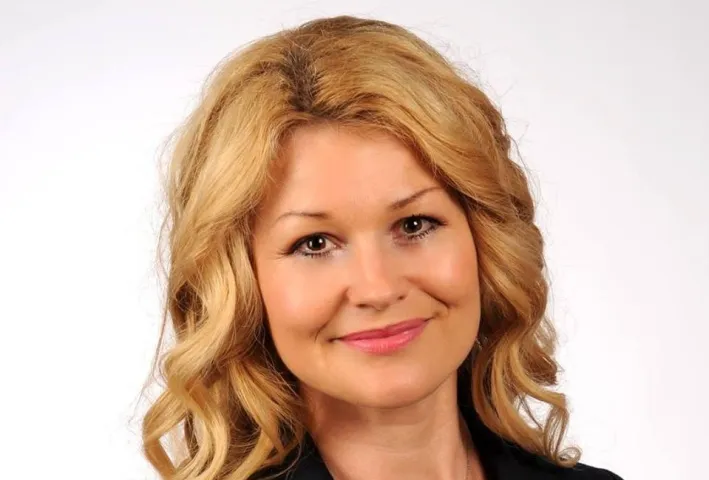 Portret uśmiechniętej kobiety o blond włosach na jasnym tle.