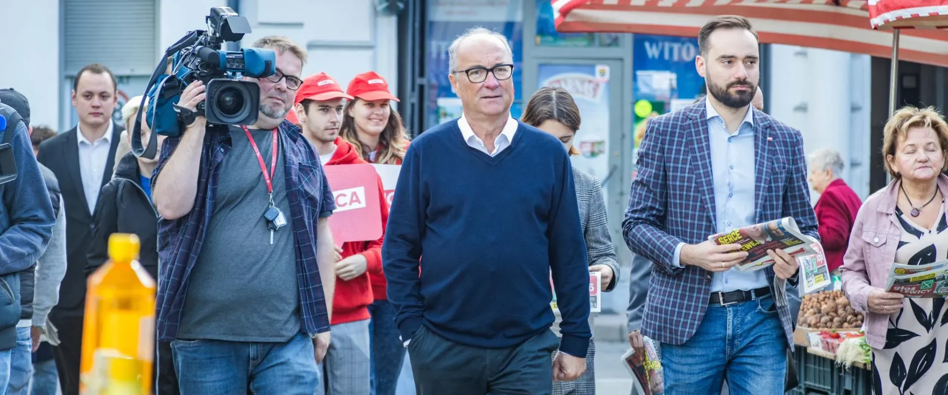 Grupa osób na ulicy podczas zgromadzenia obywatelskiego związanego z referendum; reporter z kamerą po lewej, w centrum stoi mężczyzna w granatowym swetrze, a po prawej trzyma gazetę.