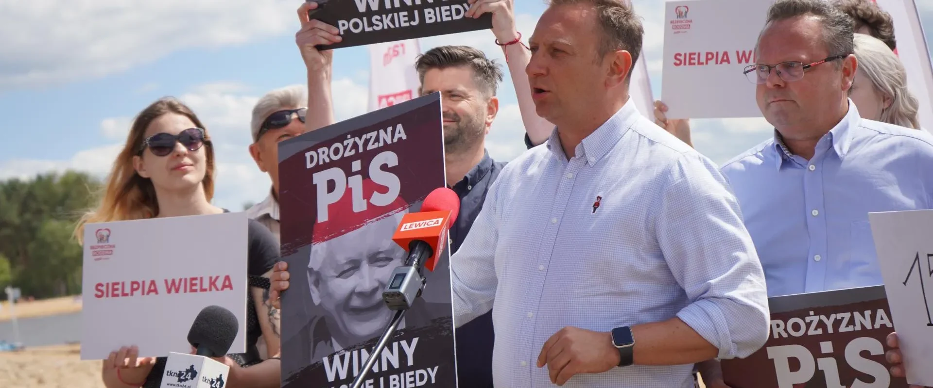 Na zdjęciu przedstawiono protest, podczas którego osoby trzymają transparenty z hasłami krytykującymi rządzących, a na pierwszym planie przemawia mężczyzna przed mikrofonem.