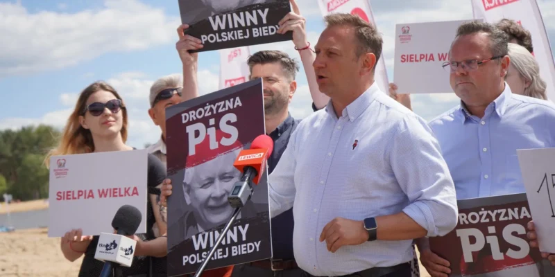 Na zdjęciu przedstawiono protest, podczas którego osoby trzymają transparenty z hasłami krytykującymi rządzących, a na pierwszym planie przemawia mężczyzna przed mikrofonem.