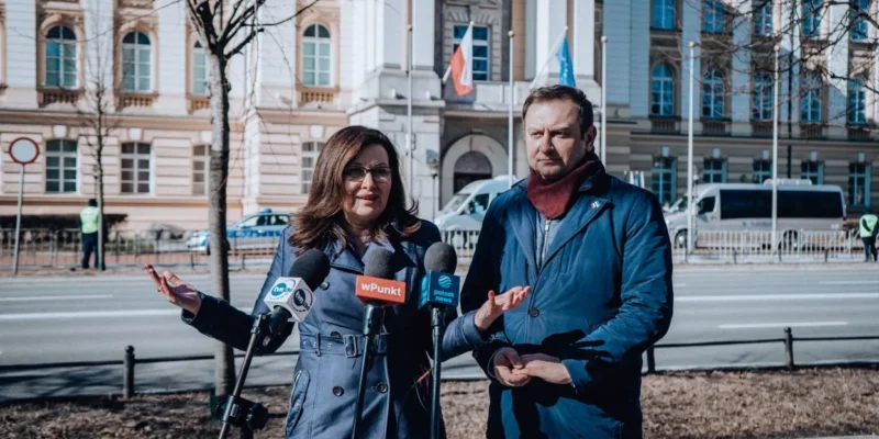 Na zdjęciu widać dwóch mówiących o aktualnych sprawach publicznych polityków, stojących przed budynkiem w słoneczny dzień.