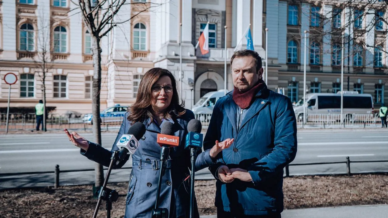 Na zdjęciu widać dwóch mówiących o aktualnych sprawach publicznych polityków, stojących przed budynkiem w słoneczny dzień.