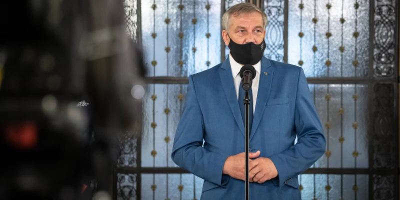 Polityk w niebieskim garniturze i czarnej maseczce stoi przy mikrofonie podczas konferencji prasowej, na tle ozdobnych żelaznych drzwi.