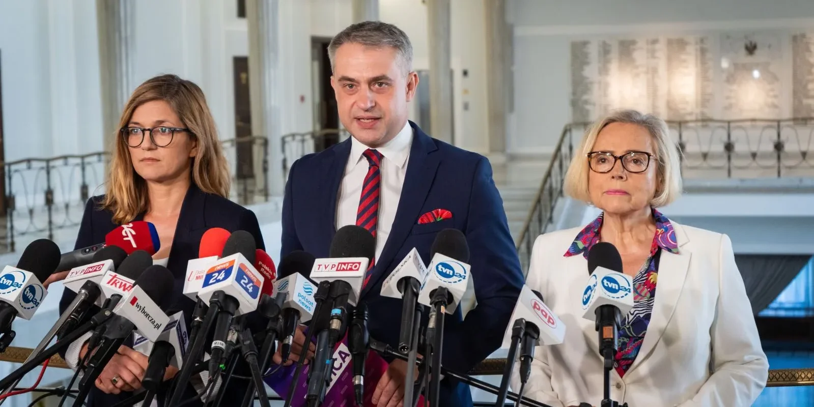 Trzy osoby stoją na konferencji prasowej w holu, przed licznymi mikrofonami; centralnie mężczyzna w granatowym garniturze z czerwonym krawatem i poszetką, po bokach kobiety w formalnych marynarkach.