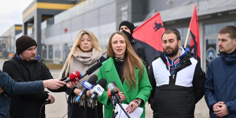 Grupa osób w różnych strojach, w tym jedna w zielonym płaszczu, prowadzi konferencję prasową na tle budynku, trzymając mikrofony i flagi.