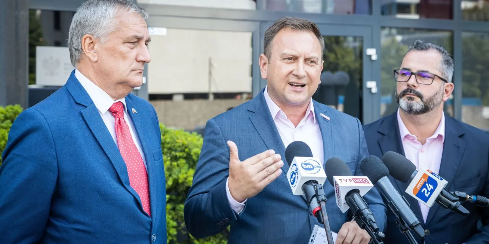 Na zdjęciu trzech mężczyzn w garniturach wygłasza oświadczenie przed kamerami i mikrofonami, stojąc obok siebie na tle budynku.