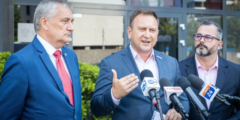 Na zdjęciu trzech mężczyzn w garniturach wygłasza oświadczenie przed kamerami i mikrofonami, stojąc obok siebie na tle budynku.