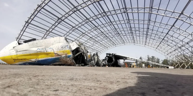 Zniszczony samolot pod ogromną metalową konstrukcją, otoczony przez resztki innego sprzętu lotniczego.