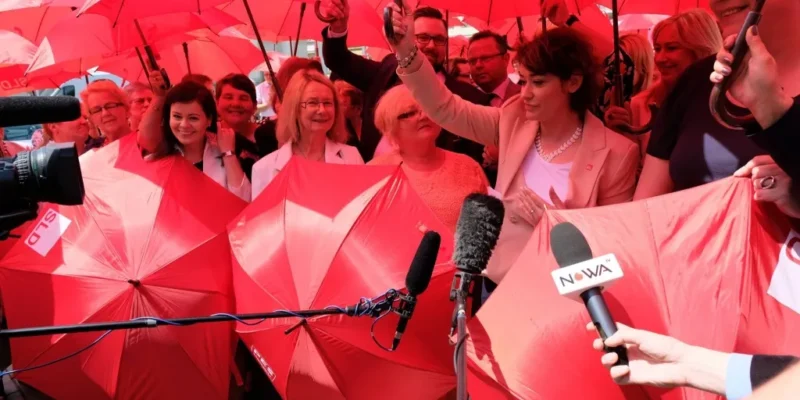 Grupa osób trzymających czerwone parasole podczas konferencji prasowej, z mikrofonami i kamerami skierowanymi ku nim.