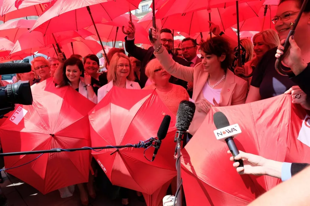 Grupa osób trzymających czerwone parasole podczas konferencji prasowej, z mikrofonami i kamerami skierowanymi ku nim.