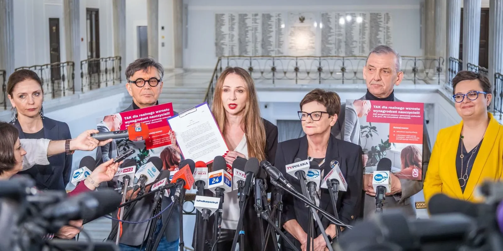 Grupa działaczy ZNP i przedstawicieli demokratycznej opozycji podczas konferencji prasowej promującej 20-procentową podwyżkę dla nauczycieli, w otoczeniu licznych mikrofonów i plakatów.