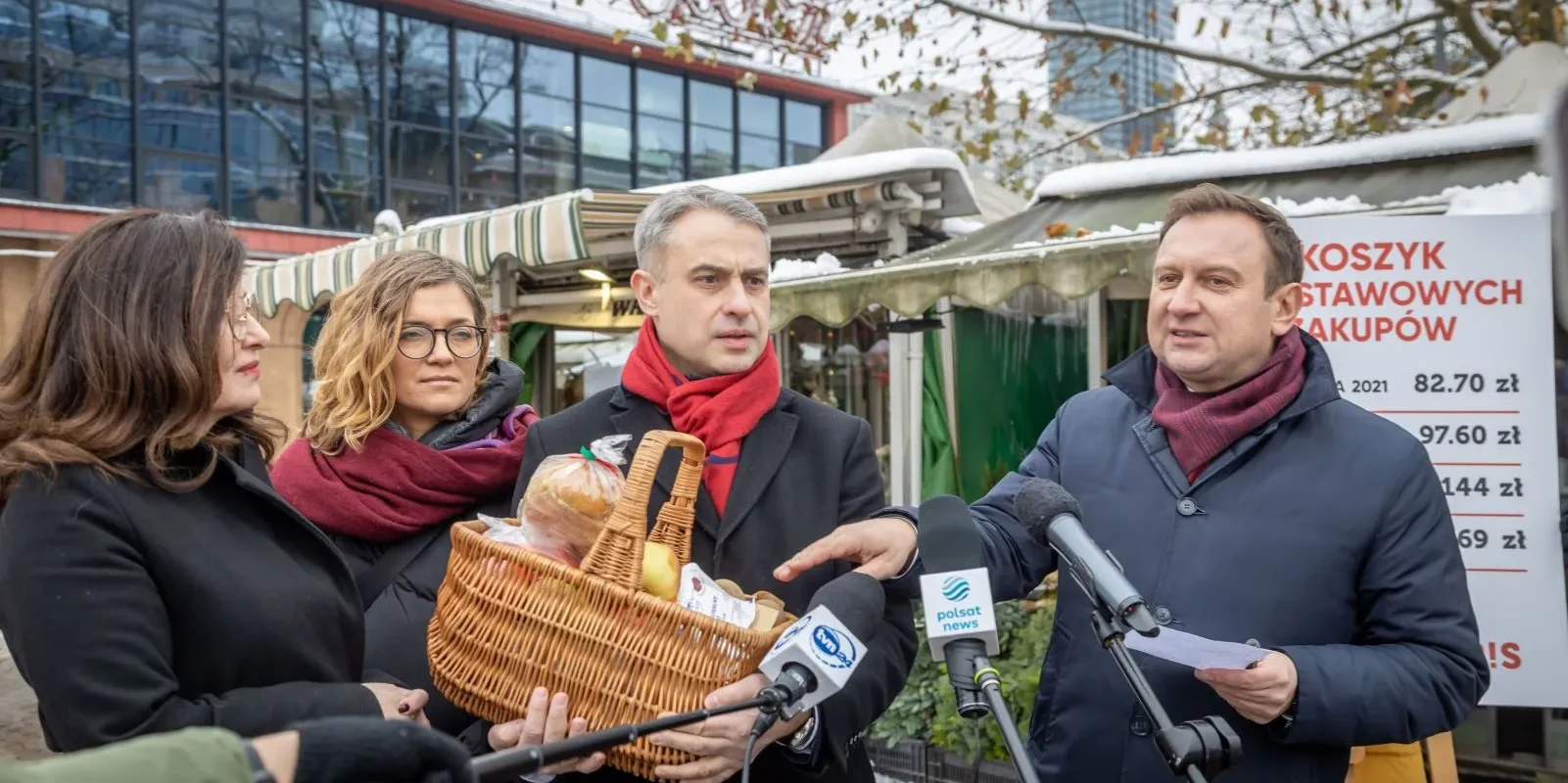 Grupa osób w zimowych ubraniach na targowisku prezentuje kosz pełen pieczywa i artykułów spożywczych podczas konferencji prasowej, obok mikrofonów i szyldu z cenami w tle.