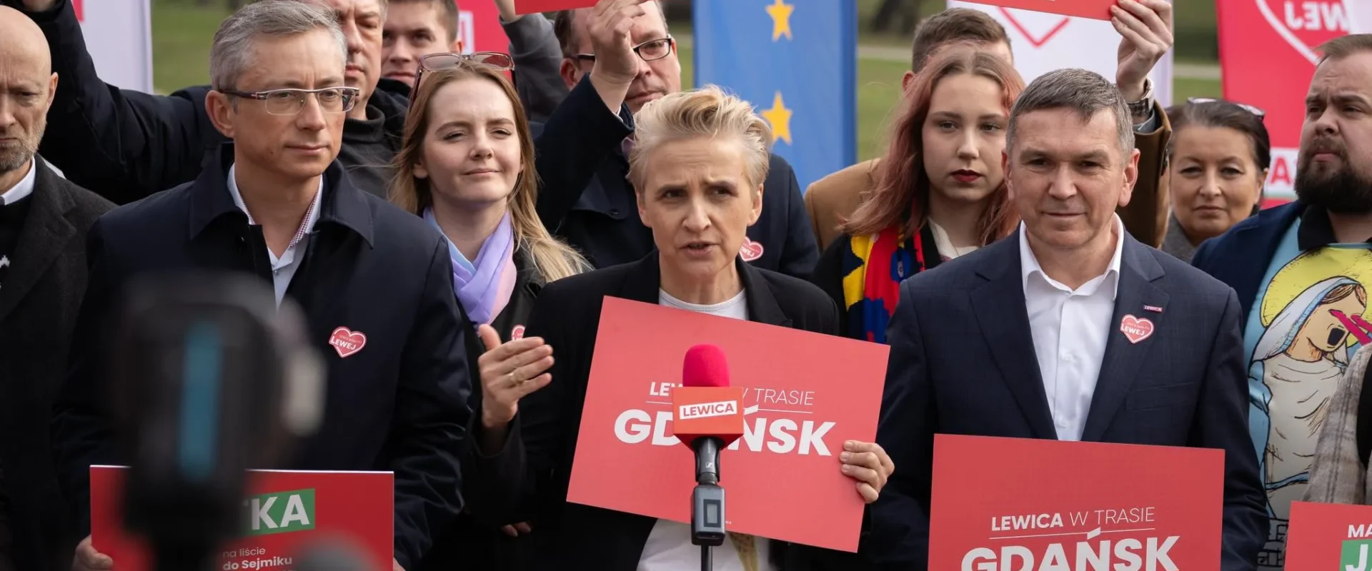 Kobieta przemawia do mikrofonu podczas wiecu wyborczego Lewicy w Gdańsku, otoczona uczestnikami trzymającymi czerwone plakaty z napisem GDAŃSK.