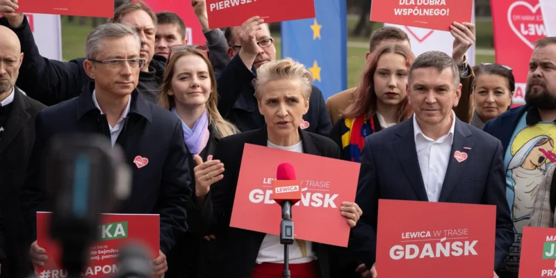 Kobieta przemawia do mikrofonu podczas wiecu wyborczego Lewicy w Gdańsku, otoczona uczestnikami trzymającymi czerwone plakaty z napisem GDAŃSK.