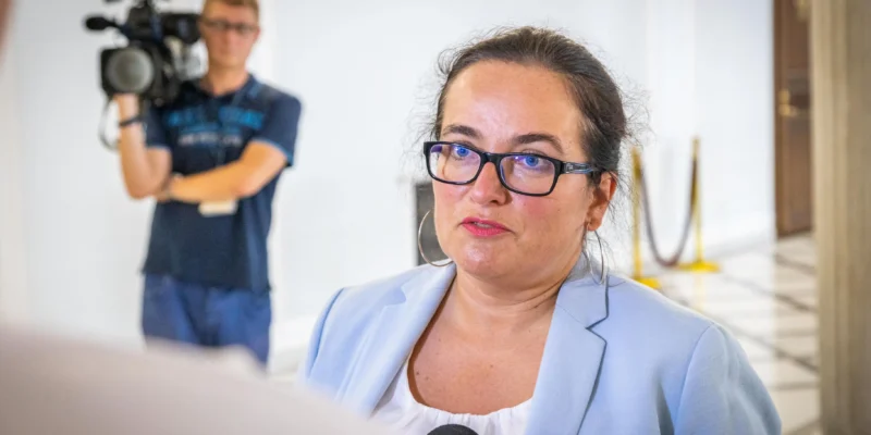 Kobieta w jasnoniebieskim żakiecie i okularach mówi do mikrofonu podczas konferencji prasowej, w tle operator kamery.