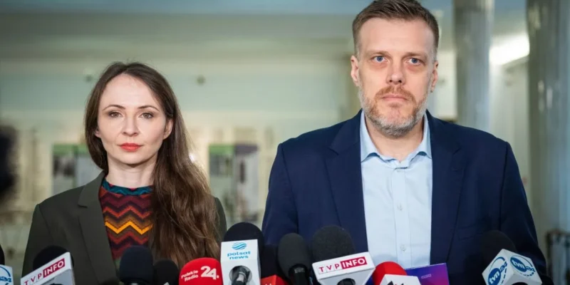 Na zdjęciu przedstawiono dwoje mówców stojących za mikrofonami, w tle widać jasne wnętrze budynku.