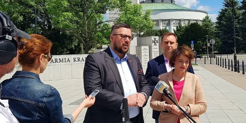 Trzy osoby podczas konferencji prasowej przed budynkiem parlamentu mówią do mikrofonów, a dziennikarze nagrywają ich wypowiedzi.
