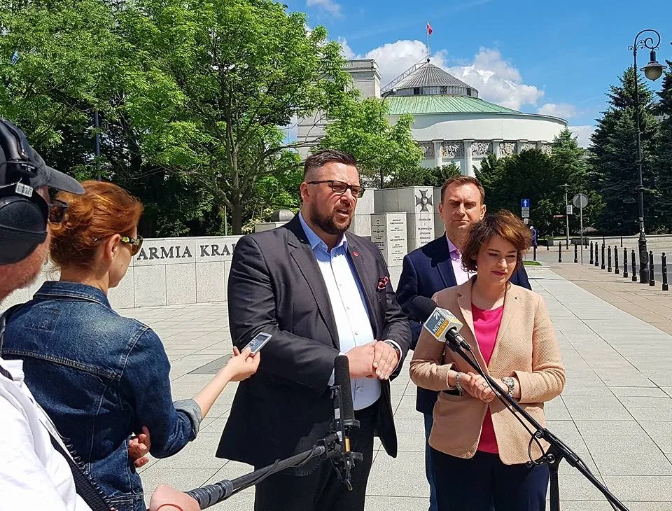 Trzy osoby podczas konferencji prasowej przed budynkiem parlamentu mówią do mikrofonów, a dziennikarze nagrywają ich wypowiedzi.