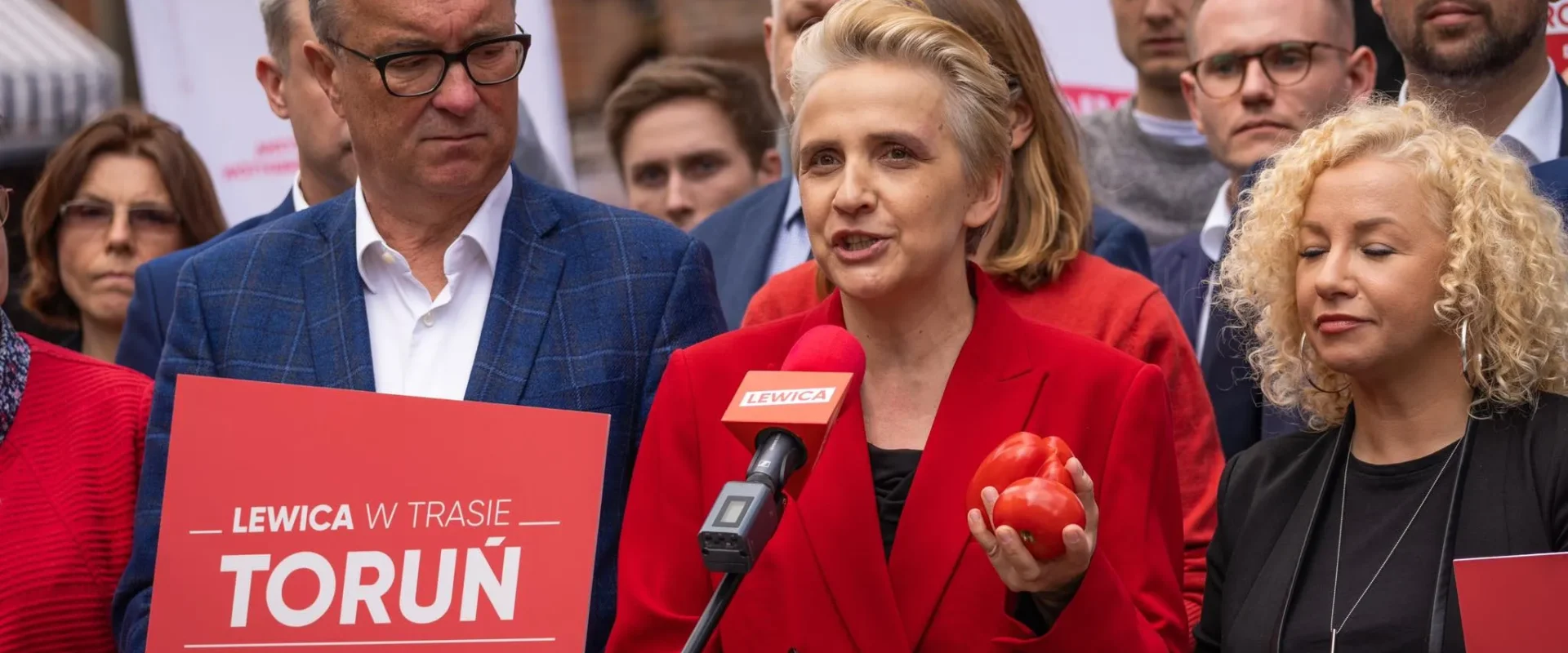 Kobieta w czerwonej marynarce przemawia do mikrofonu z logo Lewicy, trzymając czerwone pomidory, otoczona przez innych uczestników i plakaty partii.