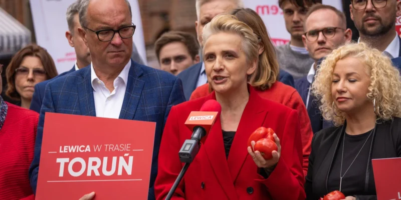 Kobieta w czerwonej marynarce przemawia do mikrofonu z logo Lewicy, trzymając czerwone pomidory, otoczona przez innych uczestników i plakaty partii.