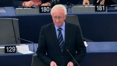 Mężczyzna w garniturze i krawacie przemawia przy mównicy na sali obrad z widocznymi numerami miejsc.