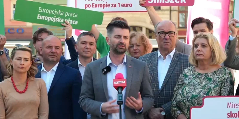 Grupa osób podczas konferencji prasowej Lewicy, z mówcą przy mikrofonie i plakatami promującymi wakacyjne bezpłatne podróże koleją dla osób do 26. roku życia.
