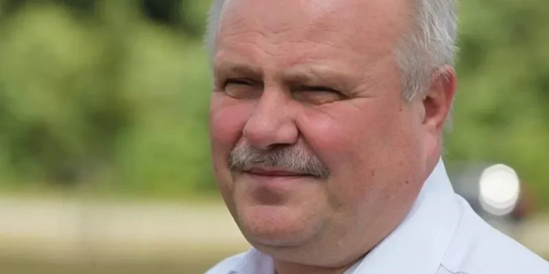 Portret starszego mężczyzny z siwymi włosami i wąsami, ubrany w białą koszulę, stojącego na tle zieleni i jeziora.