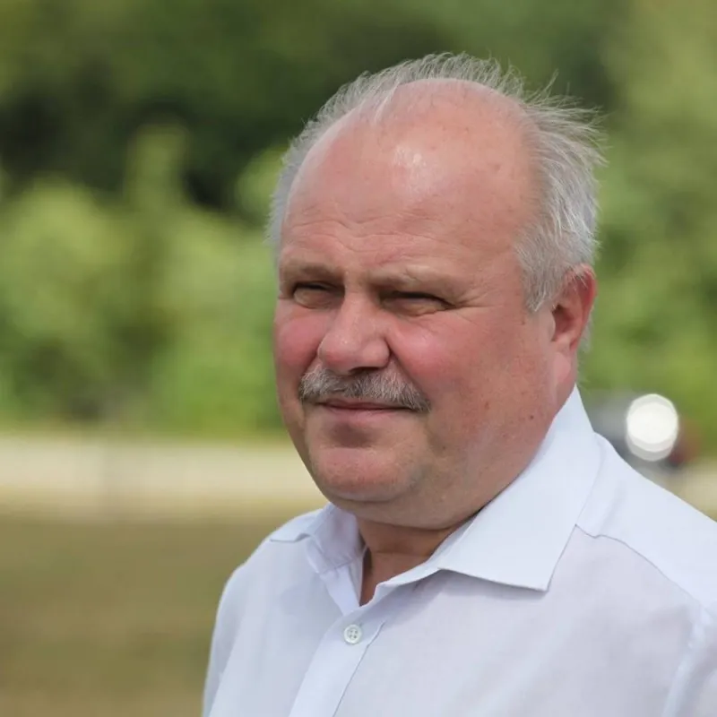 Portret starszego mężczyzny z siwymi włosami i wąsami, ubrany w białą koszulę, stojącego na tle zieleni i jeziora.