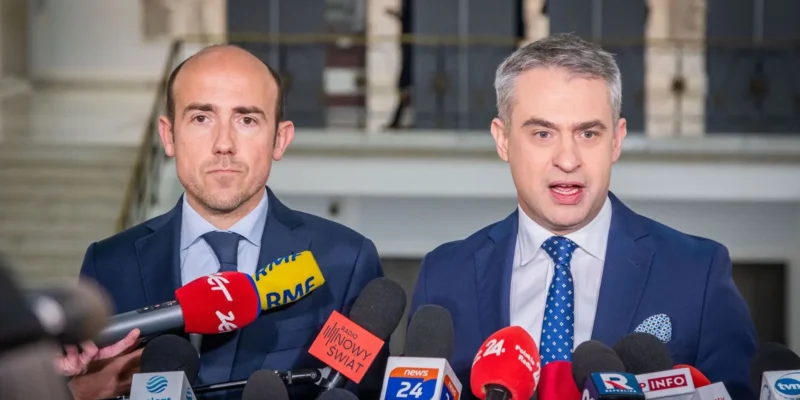 Na zdjęciu dwóch mężczyzn stoi przed mikrofonami podczas konferencji prasowej, wyrażając swoje myśli na temat aktualnych wydarzeń.