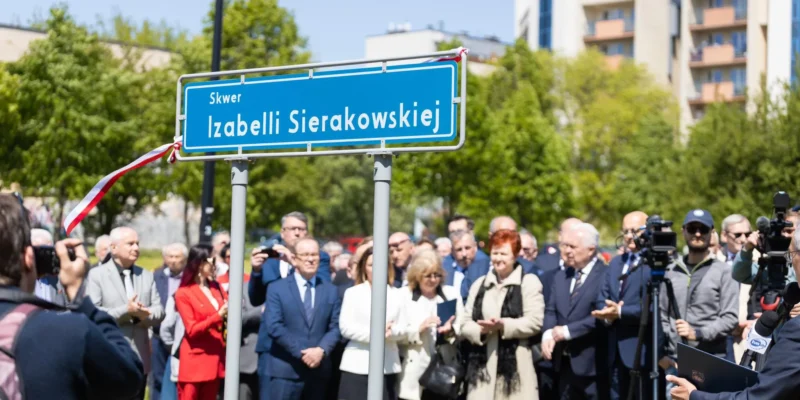 Na zdjęciu znajduje się uroczyste odsłonięcie tablicy z nazwą skweru Izabelli Sierakowskiej, podczas którego mieszkańcy oraz zaproszeni goście obecni są w tle.