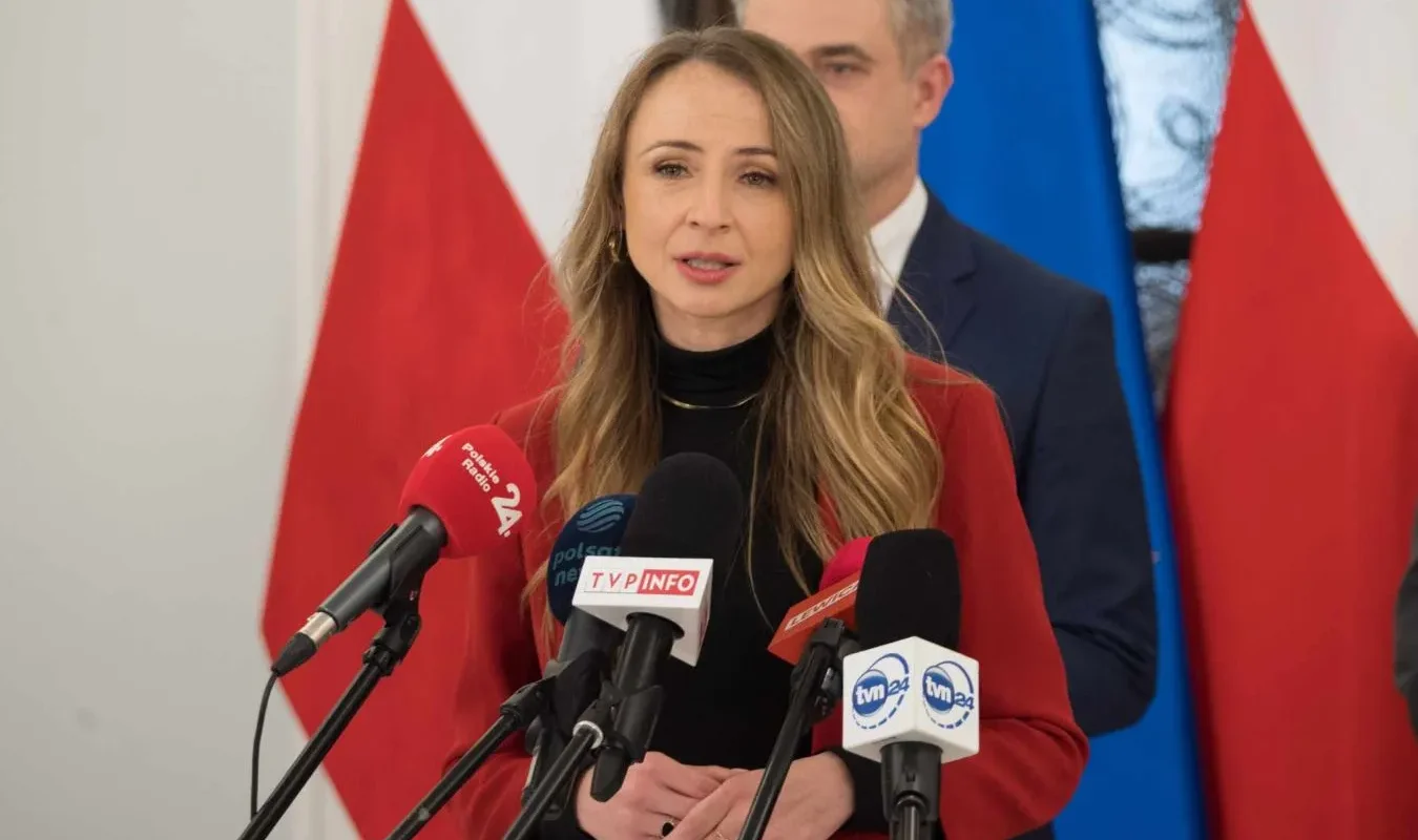 Kobieta w czerwonej marynarce przemawia do mediów podczas konferencji prasowej, stojąc przed mikrofonami z logotypami stacji telewizyjnych i radiowych, na tle flag państwowych.