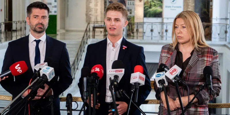 Trzy osoby w garniturach stoją przed zestawem mikrofonów podczas konferencji prasowej w dużym holu.