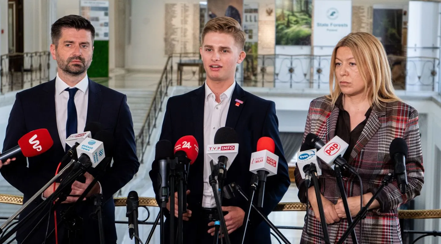 Trzy osoby w garniturach stoją przed zestawem mikrofonów podczas konferencji prasowej w dużym holu.