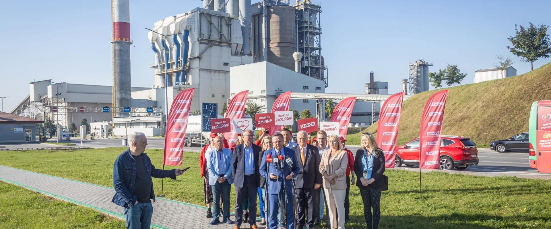 Grupa polityków Lewicy wygłasza konferencję prasową przed zakładem przemysłowym, z czerwonymi flagami Lewicy na tle instalacji.