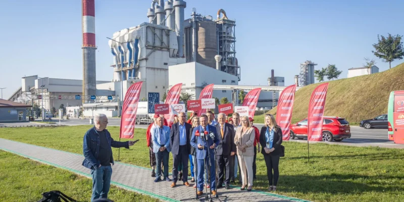 Grupa polityków Lewicy wygłasza konferencję prasową przed zakładem przemysłowym, z czerwonymi flagami Lewicy na tle instalacji.