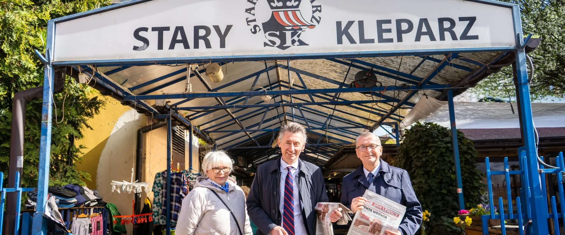 Na zdjęciu widać grupę trzech osób stojących przed budynkiem z napisem "STARY KLEPARZ", trzymających gazetę.