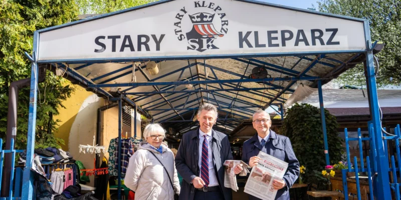 Na zdjęciu widać grupę trzech osób stojących przed budynkiem z napisem "STARY KLEPARZ", trzymających gazetę.