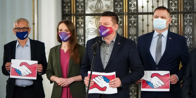 Czterech dorosłych w garniturach i marynarkach stoi na konferencji prasowej, trzyma plakietki z wizerunkiem uścisku dłoni, wszyscy mają maseczki, a za nimi ozdobna metalowa brama.