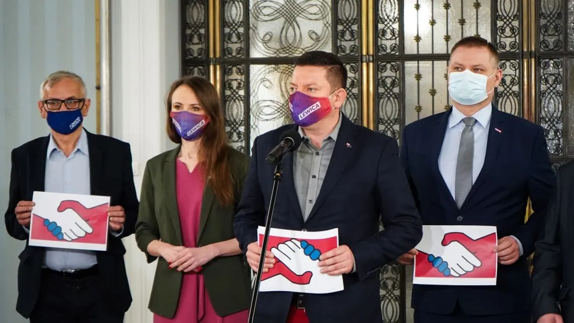 Czterech dorosłych w garniturach i marynarkach stoi na konferencji prasowej, trzyma plakietki z wizerunkiem uścisku dłoni, wszyscy mają maseczki, a za nimi ozdobna metalowa brama.